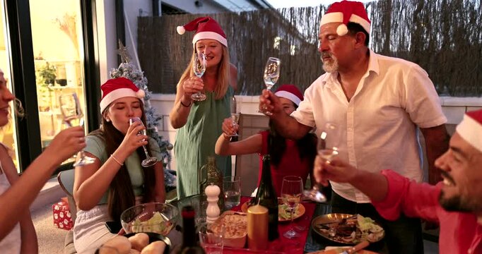 Multigenerational family wearing santa hats clinking champagne glasses and toasting during a festive christmas eve dinner party on an outdoor patio, celebrating together with joy