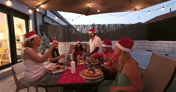 Happy multi generation family wearing santa hats enjoying a festive christmas dinner on a decorated patio, toasting with wine glasses under warm string lights during a beautiful summer evening