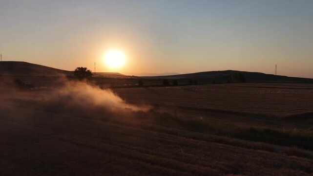 Aerial drone shot of a heavy machinery combine harvester cutting ripe wheat in a field, kicking up clouds of dust at sunset.