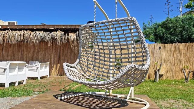 Hanging wicker chair sways gently in a sunny outdoor setting with a wooden deck, surrounded by grass and a rustic fence, creating a serene relaxation space