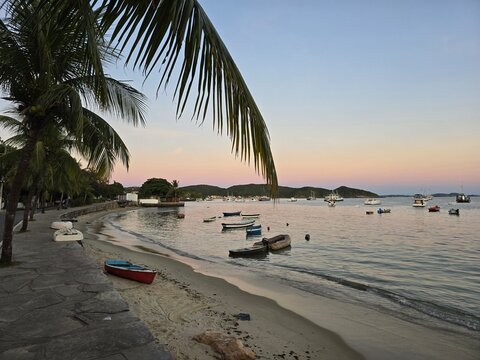 Peaceful morning at Orla Bardot with palm leaf framing, red wooden boat on the beach and fishing boats at dawn in Armacao dos Buzios, Rio de Janeiro, Brazil.