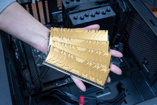 Four gold DDR memory sticks held in technician's hand, against background of open case of high-end gaming PC or graphics workstation. Concept of rising prices for RAM