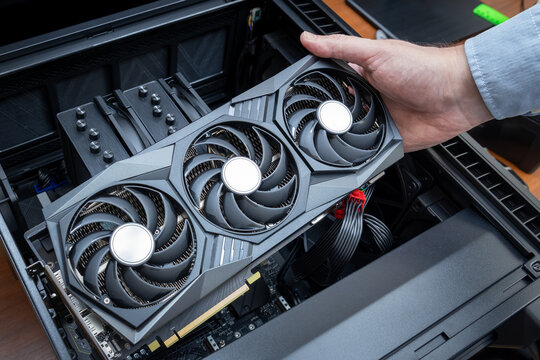 A technician installs a powerful triple-fan gaming graphics card on a gaming PC motherboard. A dual-tower CPU cooler and black power cables are visible