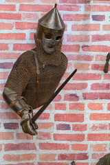 Obraz premium Medieval knight in chainmail stands against brick wall. Helmet with pointed top covers his face. He grips a sword with both hands. Brick wall behind him is textured and red