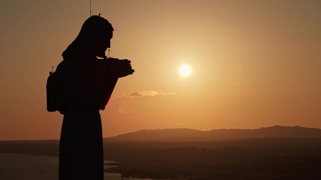 Dramatic silhouette of the Cristo Rei monument appearing to hold the setting sun above Almada, Portugal, with a warm orange sky and distant hills.