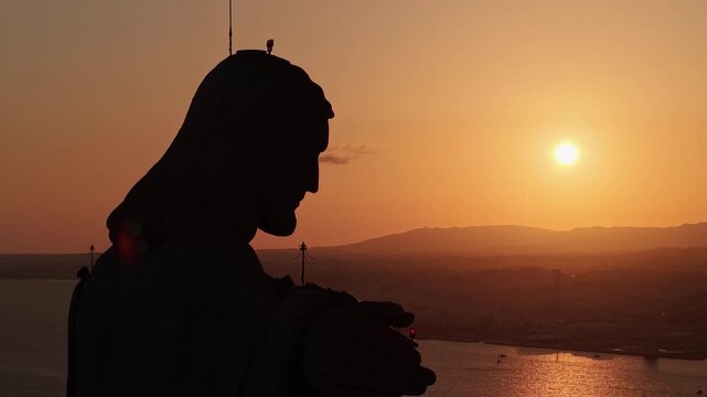 Close aerial view of the Cristo Rei monument in silhouette against a glowing orange sunset above Almada and the Tagus estuary in Portugal.