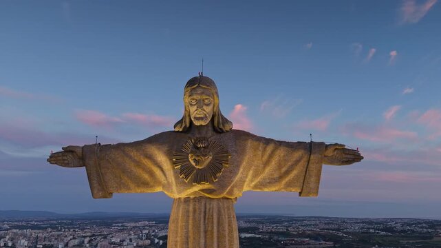 Close frontal aerial view of the illuminated Cristo Rei monument above Almada, Portugal, beneath a blue twilight sky with soft pink clouds.
