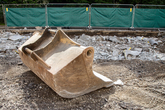 An excavator bucket positioned on a construction site. Is situated behind a wire mesh fence .The ground surrounding the bucket consists of dirt and debris. Rusty attachment are to a digging, grading. 