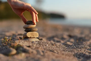 Fotobehang Rustgevend Woman lifestyle concept detail where a hand balances smooth zen stones on a sandy beach. Peaceful outdoor meditation scene at sunset light creating a calm and mindful atmosphere.  © SHOTPRIME STUDIO