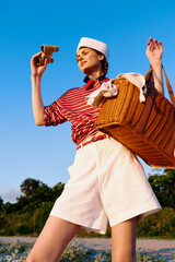 Young woman wearing a sailor hat and striped shirt holds vintage binoculars and a picnic basket on a sunny beach. The summer leisure theme captures an authentic travel adventure. © SHOTPRIME STUDIO