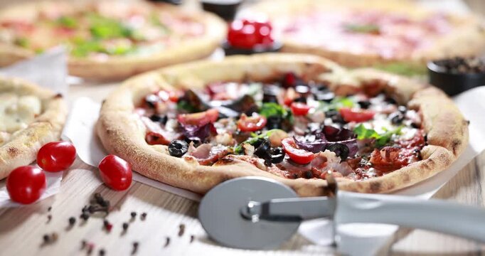 Different yummy pizzas, ingredients and cutter on wooden table, closeup. Camera moving