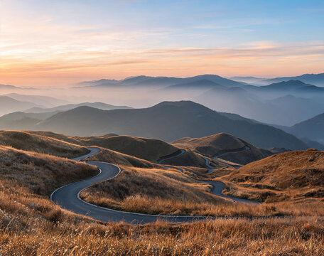 A winding asphalt road snakes through dry grassy rolling hills, with layered misty mountains under a soft sunrise sky.