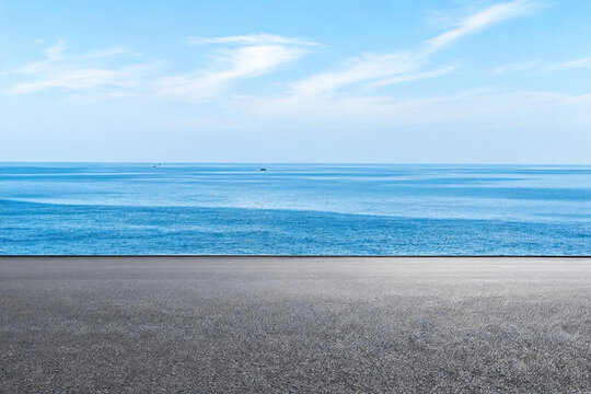 An empty flat asphalt platform stretches toward a calm blue sea under a bright clear sky, with distant vessels on the water.