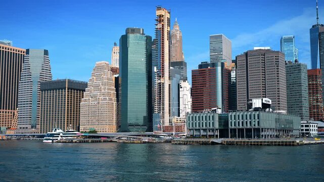 Lower Manhattan skyline above East River waterfront. A ferry and docked sailing vessel sit below dense towers in clear Independence Day light.