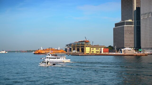 Boats passing South Ferry waterfront in Lower Manhattan. A white tour vessel and small patrol craft leave wakes beside the orange Staten Island Ferry.