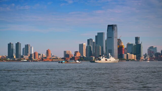 Naval frigate dressed with signal flags passing Jersey City skyline. A tugboat pushes a barge across the water beneath modern high rise towers at dawn.