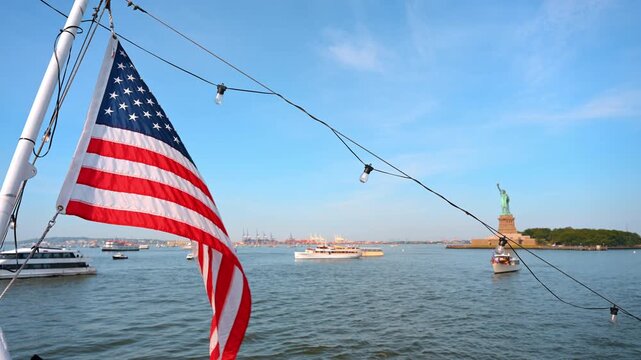 American flag flying above New York Harbor near Statue of Liberty. String lights and boats drift near Liberty Island beneath a bright afternoon sky.