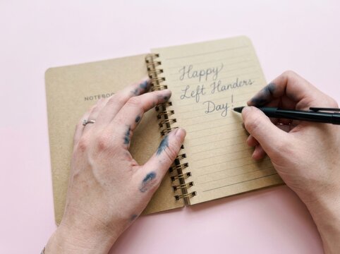 Closeup of a lefthanded person writing happy left handers day in a spiral notebook with inkstained fingers on a pink background