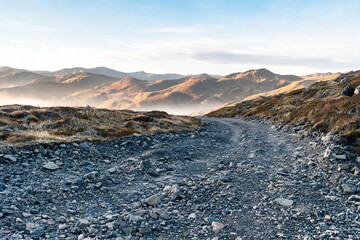 Fototapeta premium A rugged rocky trail winds through highland terrain, leading toward hazy mountain ranges under a clear sunlit sky.