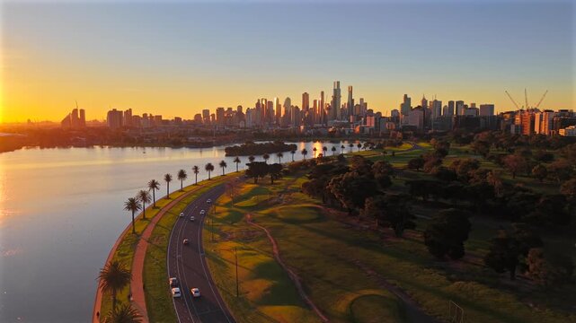 Low aerial drone video flying toward Melbourne CBD at sunset over Albert Park Lake, showing palm-lined roads, golf course, Southbank and golden city skyline