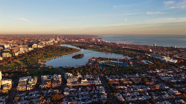 Aerial drone video facing south over Albert Park Lake in Melbourne, showing parkland, city suburbs, golden light and the Port Phillip Bay coastline