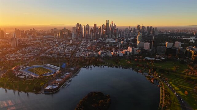 Aerial drone video flying backward at sunset over Albert Park Lake, revealing South Melbourne, Southbank and the Melbourne CBD skyline in golden evening light