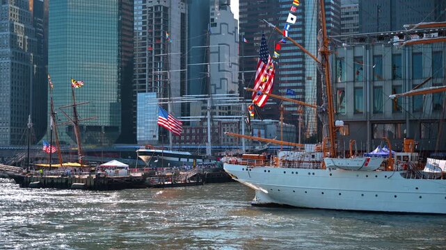 U.S. Coast Guard tall ship with signal flags in Lower Manhattan. A white hull and orange rigging fill the waterfront beside crowded piers and glass towers.