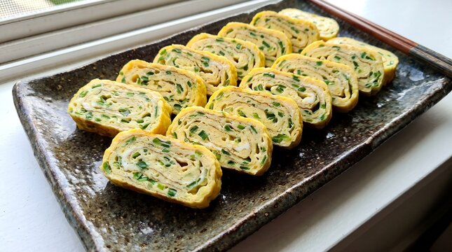 Slices of Gyeran-mari (rolled omelet) arranged neatly on a dark ceramic rectangular plate, showing the layers and green onion inclusions.