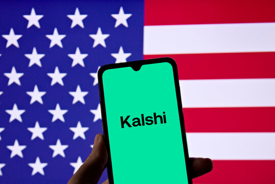 Smartphone displaying the Kalshi prediction market platform logo on a green screen against a United States flag backdrop.