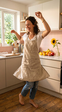 Vertical photo of a smiling barefoot woman dancing in a kitchen. Happy female wearing a linen apron having fun at home. Spontaneous joy and playful living concept
