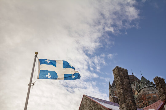 Quebec flag with fleur-de-lys and white cross waving against a winter sky, with Chateau de Frontenac in the background above historic stone buildings in Old Quebec.