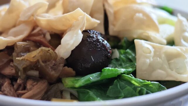 Slow motion camera view of a full bowl of Indonesian chicken noodles with fried wontons, shiitake mushroom, chicken meat, and green vegetables.
