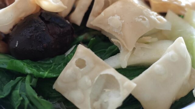 Detailed macro slow motion shot of chicken noodle toppings including crispy wonton crackers, shiitake mushroom, and green mustard greens in a white bowl.

