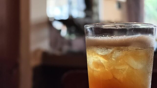 Side angle slow motion focus on the top rim and ice cubes of a cold glass of iced lemon tea in a cafe background.
