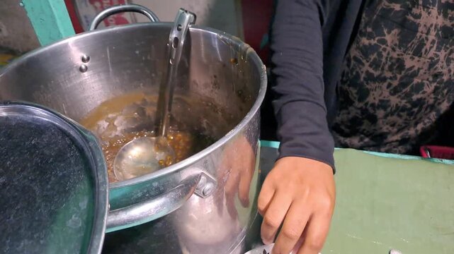 Indonesian vendor preparing Es Kacang Ijo for takeaway order. Process includes scooping mung beans into plastic, shaving ice with vintage manual machine, and pouring coconut milk. Street food cart.