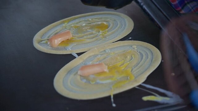 Asian street food vendor preparing thin pancake rolled crepe with sausage on hot pan.