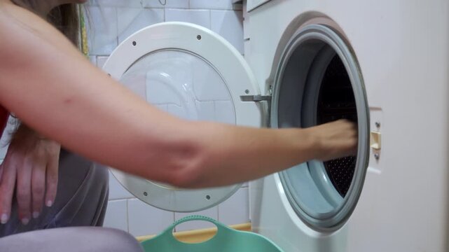 Close view of adult placing damp clothing into open washer drum inside tidy laundry room, with pale walls, plastic basket nearby, soft overhead light, and everyday domestic cleaning activity