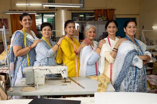 Group of skilled Indian women workers standing together inside a textile export house with their hands on each other's shoulders while looking directly at the camera. The gesture represents teamwork.