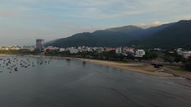Smooth drone flight following the calm shoreline of Da Nang Bay with fishing boats, mountains and the first light of sunrise.