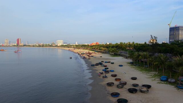 Smooth rotating aerial drone shot revealing Da Nang beach, fishing basket boats, palm-lined coastline and city skyline during sunrise. Peaceful tropical seaside scene in central Vietnam.