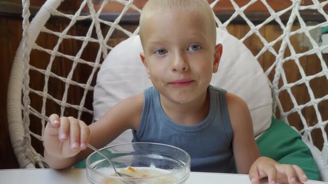 Blond preschool boy sits in woven hanging chair eating cereal from glass bowl with spoon, calm expression and direct gaze, gray sleeveless top, soft daylight, relaxed summer home setting