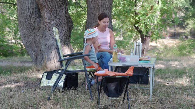 Middle-aged woman grilling sausages on small kettle barbecue under large shade trees beside folding table and plastic chairs, with cooler, condiments, paper cups, and relaxed outdoor picnic mood in