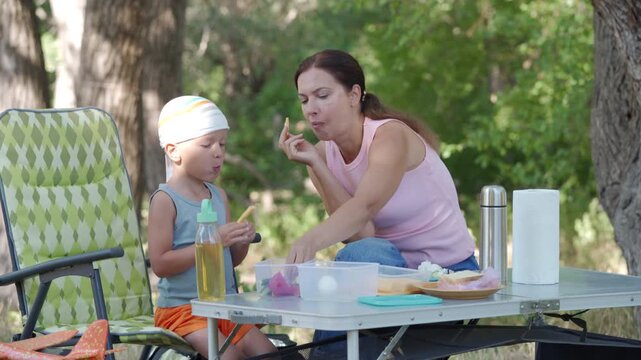 Woman feeds toddler seated at picnic table in shaded park during daytime, child in sleeveless top while woman offers food from spoon, with water bottle, juice, lunch items, and calm family