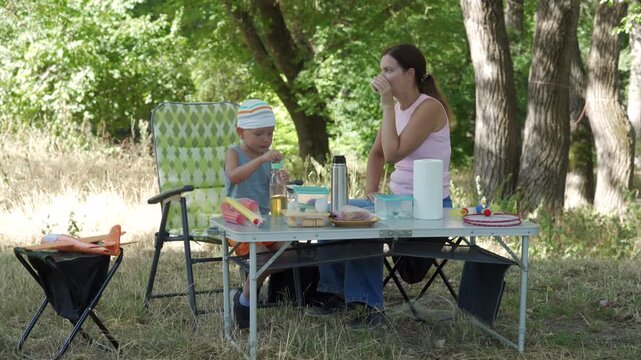 Mother and young child prepare food and set table during relaxed picnic beneath leafy trees in warm daylight, with portable grill, folding chairs and casual outdoor gathering nearby