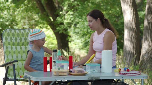 A loving mother and her happy son eat a snack together at a picnic table. They share a tender, cheerful moment during lunch. A peaceful green forest surrounds them.