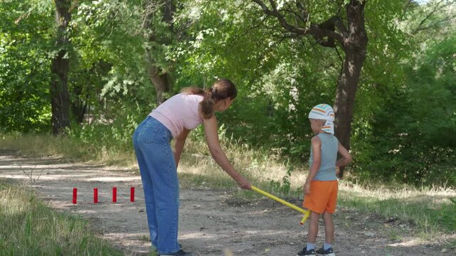 A loving mother and her happy son joyfully play a game of gorodki on a dirt path. They share a cheerful, bonding moment. A peaceful green forest surrounds them.
