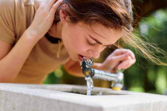 Woman drinking water from a public fountain tap outdoors, closeup portrait of hydration and refreshment. Young female leaning over stone basin with flowing clear water.