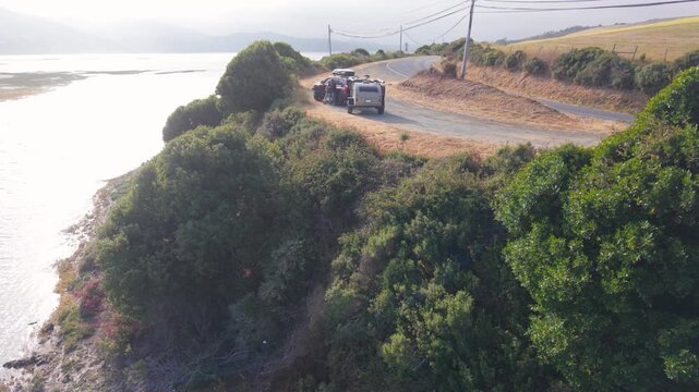 Aerial views show a teardrop camper trailer parked by the roadside in California near water as the sun sets on summer evening
