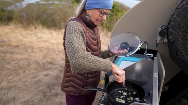A senior woman happily prepares broccoli on her teardrop camper stove while boon docking along a riverside in California.