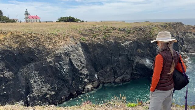 A mature woman hikes along cliffs by the Pacific Ocean toward Cabrillo Lighthouse in Mendocino California.